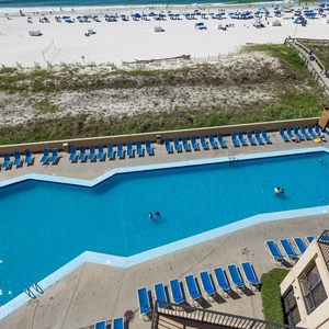 The view showcases a vibrant pool area with blue loungers and a lively beach just beyond the white sand