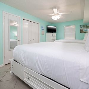 Bright turquoise walls complement two queen beds with white linens, a mounted TV, and a beach art in this inviting bedroom