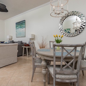 Bright seating area features a cozy sofa and armchairs, with a dining table adorned by fresh flowers and a view of the beach