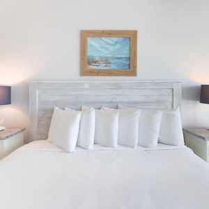 This inviting bedroom features a plush bed with numerous pillows, stylish lamps, and a serene coastal painting