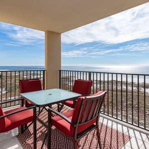 Balcony features a glass-top table with red chairs, offering a serene view of the beach and sparkling gulf waters