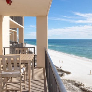 A wooden dining set on the balcony offers a serene view of the beach and tranquil blue waters, perfect for relaxation