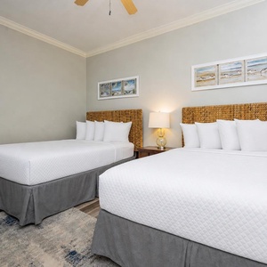 This guest bedroom features two queen beds with woven headboards, soft linens, and beach-themed artwork for a relaxing stay
