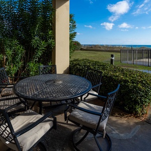 A shaded outdoor patio area features a round table surrounded by cushioned chairs, with a view of the beach beyond
