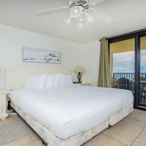 Bright master bedroom featuring a king bed, elegant lamp, and sliding glass doors leading to a balcony with a beach view