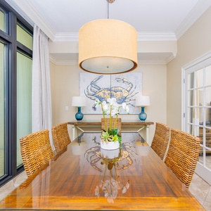 This dining area features a stylish table with woven chairs, a vibrant crab artwork, and a glimpse of the cozy bedroom beyond