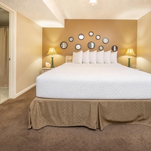 A spacious bedroom features a large king bed with multiple pillows, decorative mirrors, and soft lighting from a bedside lamp