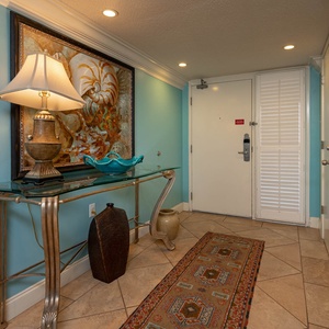 The entryway features a turquoise wall, decorative artwork, a glass console table with a lamp, and a patterned runner rug