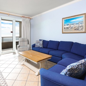 The living area features a blue sectional sofa, a wooden coffee table, and a stunning beach view through sliding glass doors