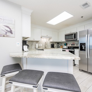 Bright, inviting kitchen features a spacious breakfast bar with seating, stainless appliances, and decorative coastal artwork