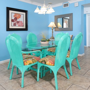 Bright turquoise dining chairs surround a glass table, complemented by floral cushions and a cheerful beach-inspired ambiance