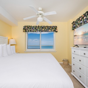 The master bedroom features a large bed, a TV, and a stunning view of the beach through bright windows
