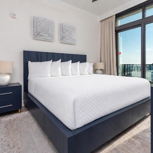 This bedroom features a stylish navy king bed, crisp white linens, and a stunning beachfront view through large windows