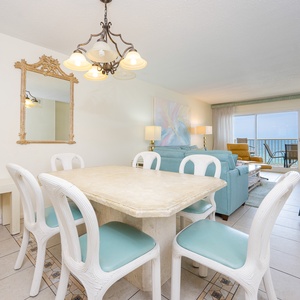 This dining area features a light stone table and white chairs, with a stunning beachfront view visible through large windows