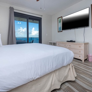 Bright master bedroom featuring a king bed, a large TV, and sliding glass doors leading to a balcony with a beach front view