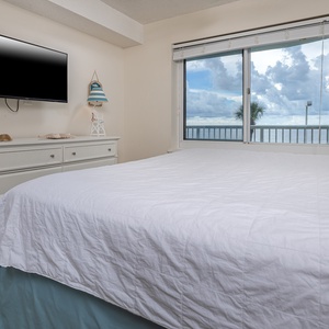 This bedroom features a large king bed, a TV, and a stunning view of the beachfront through expansive windows