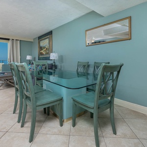 This inviting dining area features a glass-top table surrounded by teal chairs, with a breathtaking view of the beach beyond