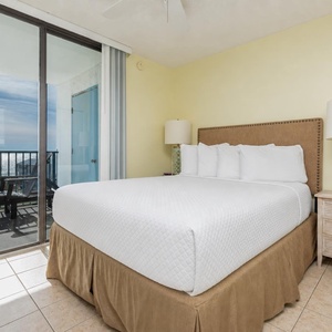 The master bedroom features a plush queen bed, a stylish lamp, and a stunning beach view from the sliding glass door