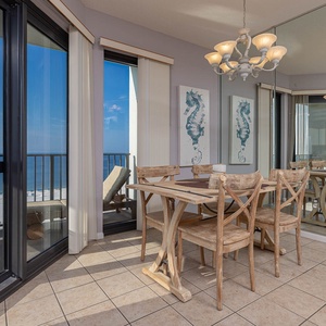 Dining area features a wooden table with four chairs, large windows showcasing a beachfront view, and coastal decor