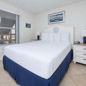 The bedroom features a bed with white linens, beach-themed decor, and a window allowing natural light to brighten the space