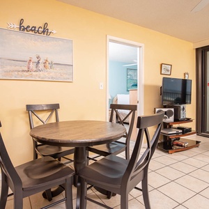 This dining area features a round table, black chairs, and a TV, with a bright beach-themed painting on the wall