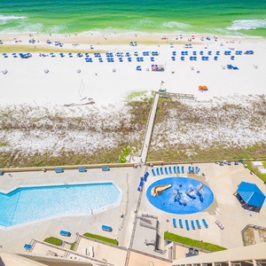 The view showcases a vibrant pool and splash play area, leading to a pristine beach with soft white sand