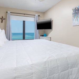 The master bedroom features a plush bed, a wall-mounted TV, and stunning beachfront views through large windows