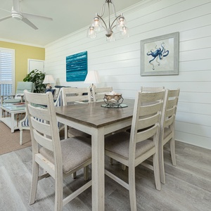The dining area features a rustic table with six chairs, complemented by coastal decor and natural light from the balcony