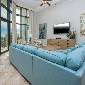 This tranquil living area features a large TV, a blue couch, and tropical decor, with a stunning beachfront balcony view