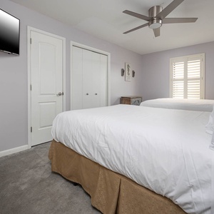 This guest bedroom features two queen beds with white linens, a ceiling fan, and a TV, creating a relaxing atmosphere