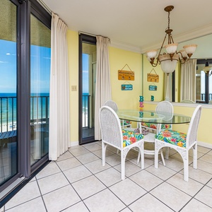 Dining area with a glass table, colorful cushioned chairs, and sliding doors leading to a balcony with a beach front view