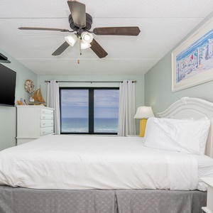 This bedroom features a large window with a stunning beachfront view, a comfy bed, ceiling fan, and a large mounted TV