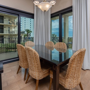 Dining area features a glass-top table surrounded by woven chairs, with large windows offering a glimpse of the beach