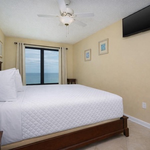 This bedroom features a king-size bed, a wall-mounted TV, and a stunning beachfront view through large windows