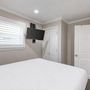This cozy bedroom features a mounted TV, light-colored walls, and a window with blinds for privacy