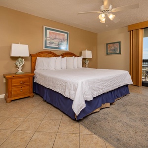 Spacious master suite featuring a king bed, warm decor, and sliding glass doors leading to a balcony with a view of the beach