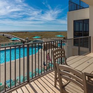 Balcony with a round table and chairs overlooks the inviting pool area and offers a glimpse of the beach in the distance
