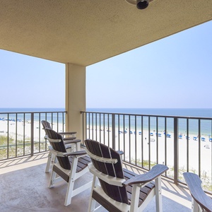 The balcony features two wooden chairs overlooking a serene beach with white sand and blue umbrellas lining the shore