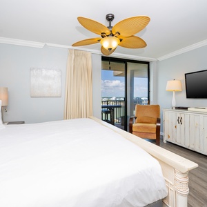 The master bedroom features a plush white bed, a large TV, and a cozy chair, with a lovely view with balcony access