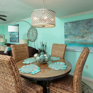 Dining area features woven chairs, a round table, and beach decor against a soothing turquoise backdrop