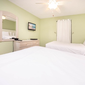 Bright green walls complement two double beds and a dresser, with a ceiling fan and a mounted TV enhancing the inviting vibe