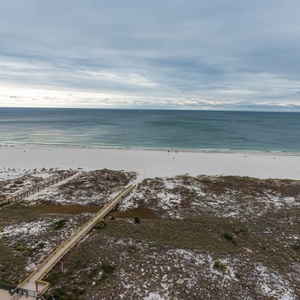 Expansive view of the beach with white sand and gentle waves, framed by lush dunes and a boardwalk leading to the shore