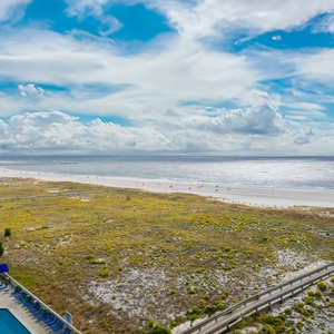 Expansive view of the beach with golden sand and gentle waves, framed by lush greenery and a sparkling pool area below