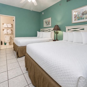 Bright teal walls complement two queen beds with white linens, a palm-themed lamp, and an en-suite bathroom with fresh towels