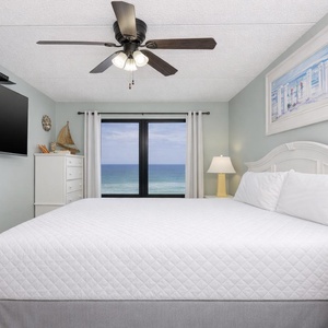 Serene master suite a large window with a stunning beachfront view, a comfy bed, ceiling fan, and a large mounted TV