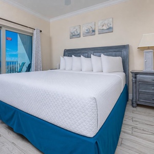 The master bedroom features a plush bed with white linens and a stunning beach view through large sliding glass doors