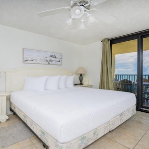 Bright master bedroom featuring a king bed, elegant lamp, and sliding glass doors leading to a balcony with a beach view