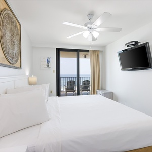 Bright master bedroom featuring a king bed, wall-mounted TV, and balcony access with an expansive view of the coastline