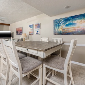 The living/dining area features a large table surrounded by white chairs, beach artwork, a TV, and queen sofa-sleeper