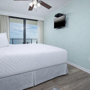 This bedroom features a plush white bed, a mounted TV, and a stunning beachfront view through large sliding glass doors